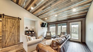 A spacious living room with a wooden ceiling, a large leather sofa, a white armchair, and a television mounted on the wall. - Cossatot River Retreat: Home w/ Fire Pit & Grill (Gillham)