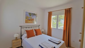 Un dormitorio con cama, dos almohadas, mesita de noche con lámpara, ventana con cortinas y un cuadro en la pared.