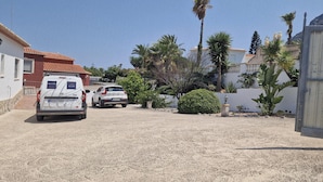 Zona residencial con una furgoneta blanca estacionada frente a una casa con techo de tejas rojas. Hay palmeras y un pequeño jardín con una fuente.