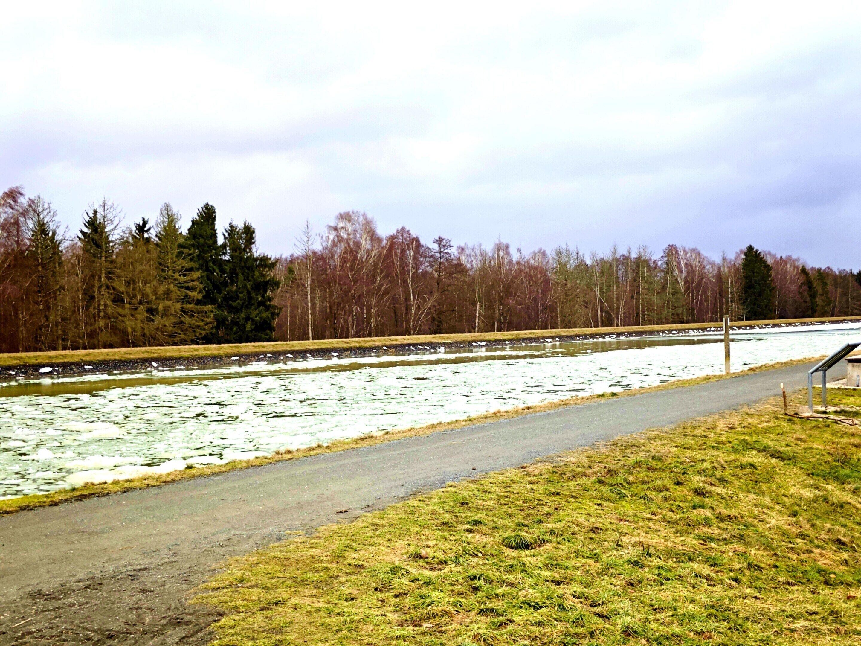 Ein Weg entlang eines zugefrorenen Gewässers mit Bäumen im Hintergrund.