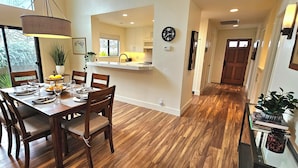 Un coin repas avec une table en bois dressée pour quatre personnes, une cuisine en arrière-plan et un couloir menant à une porte.