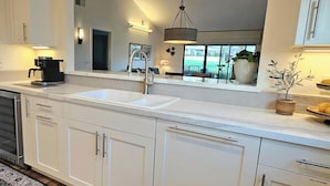 Une cuisine moderne avec des meubles blancs, un évier à deux bacs, une machine à café et une grande fenêtre donnant sur l’extérieur.