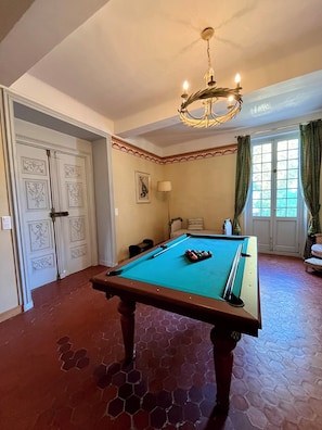 Una mesa de billar en una habitación con suelo de patrón hexagonal, una araña y una puerta con paneles decorativos.