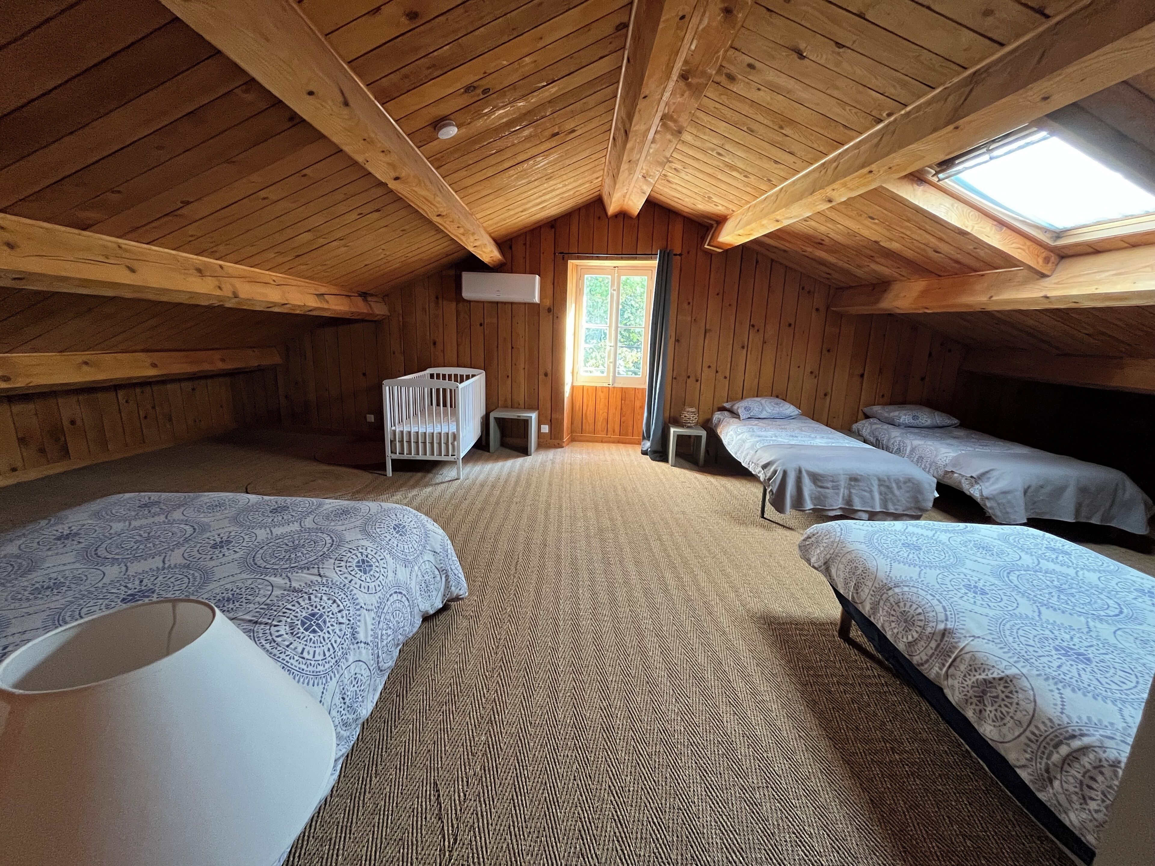 A cozy attic bedroom with wooden walls, a sloped ceiling, and a skylight. It includes two beds, a crib, and a small table.
