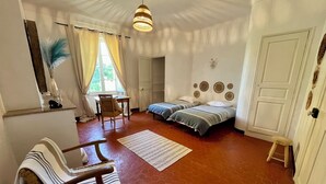 A room with two beds, a chair, a desk, a window with curtains, a hanging light fixture, and a door.