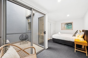 A hotel room with a bed, a chair, a small table with a lamp, a painting on the wall, and a sliding glass door leading to a balcony. - Sea Spray Apartment (Lorne)