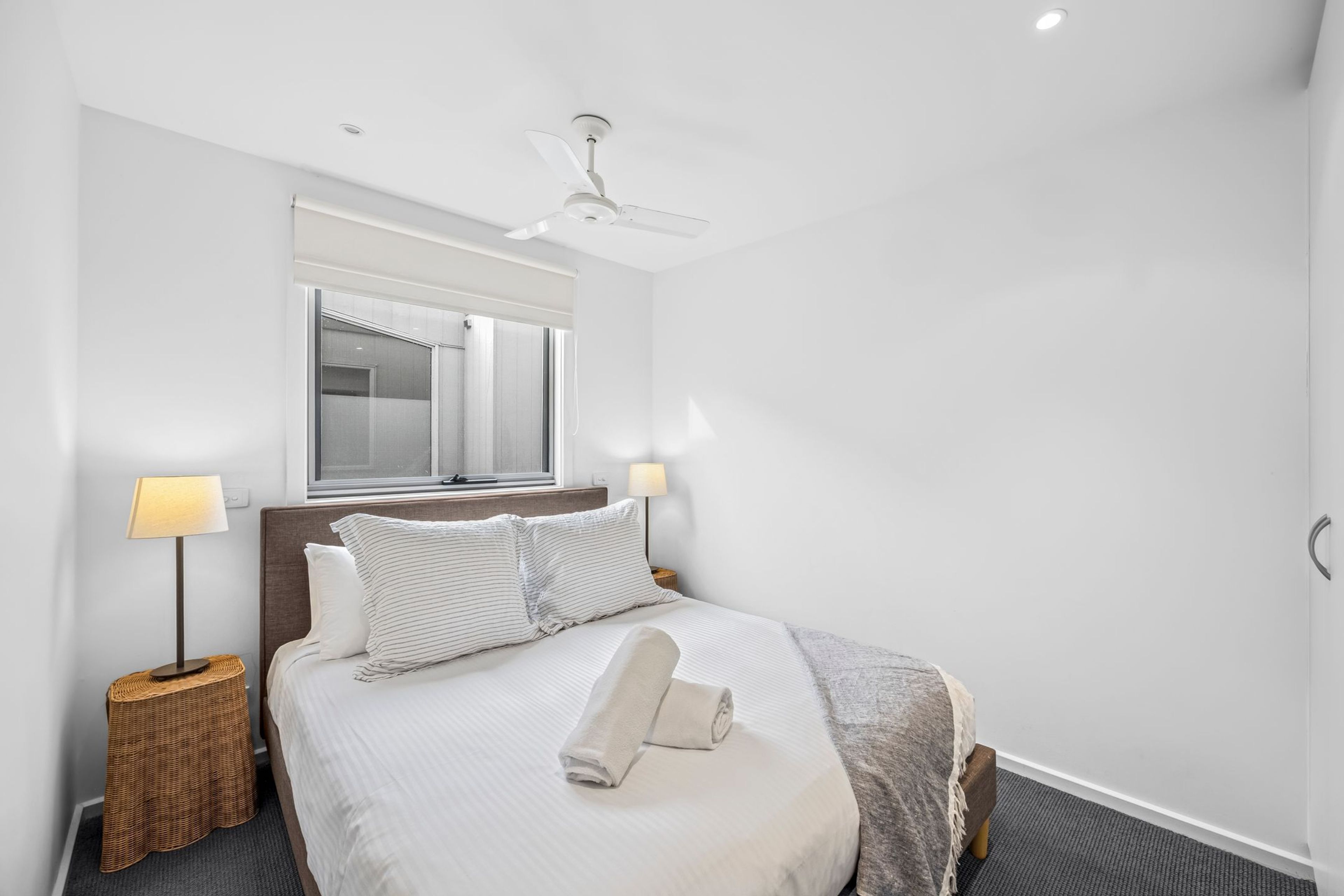 A hotel room with a bed, two bedside lamps, a ceiling fan, a window with a view, and a mirror on the wall.