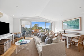 A modern living room with a large flat-screen TV, a sofa set, a dining table, and a large window offering a view of the outdoors. - Oceanview on Mountjoy (Lorne)