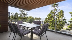 Et moderne udendørs spiseområde med et glasbord og stole, med udsigt over et naturskønt landskab med træer og vand.