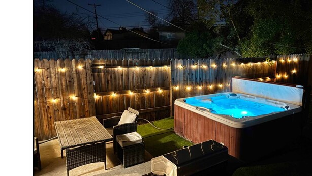 Un patio trasero con cerco de madera, luces colgantes, un jacuzzi y mobiliario exterior.