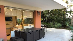 Un patio al aire libre con comedor, que incluye una mesa grande y sillas, un televisor y una cocina visible a través de puertas correderas de vidrio.