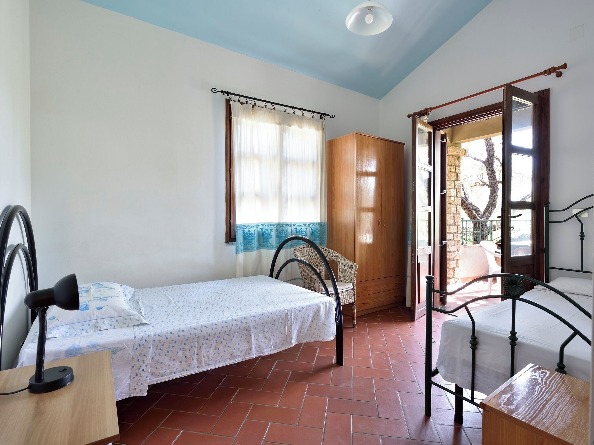A small, well-lit room with a bed, a chair, a wooden wardrobe, and a window with curtains.