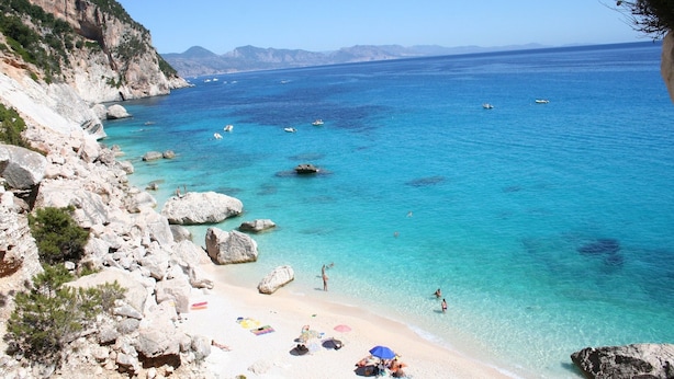 A coastal beach with clear turquoise waters, rocky terrain, and a few people enjoying the sun.