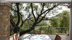 A balcony with a table covered in a tablecloth, surrounded by chairs, overlooking a garden with trees and a fence.