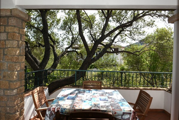 A balcony with a table covered in a tablecloth, surrounded by chairs, overlooking a garden with trees and a fence.