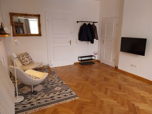 A modern living room with a white armchair, a patterned rug, a wooden herringbone floor, a mounted TV, and a coat rack with jackets. - Vacation apartment Royal Villa - Watzmannblick (Berchtesgaden)