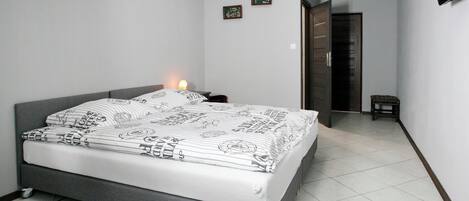 A bedroom with a grey bed, white bedding, a bedside table, a nightstand, a small stool, a door, and a wall-mounted TV.
