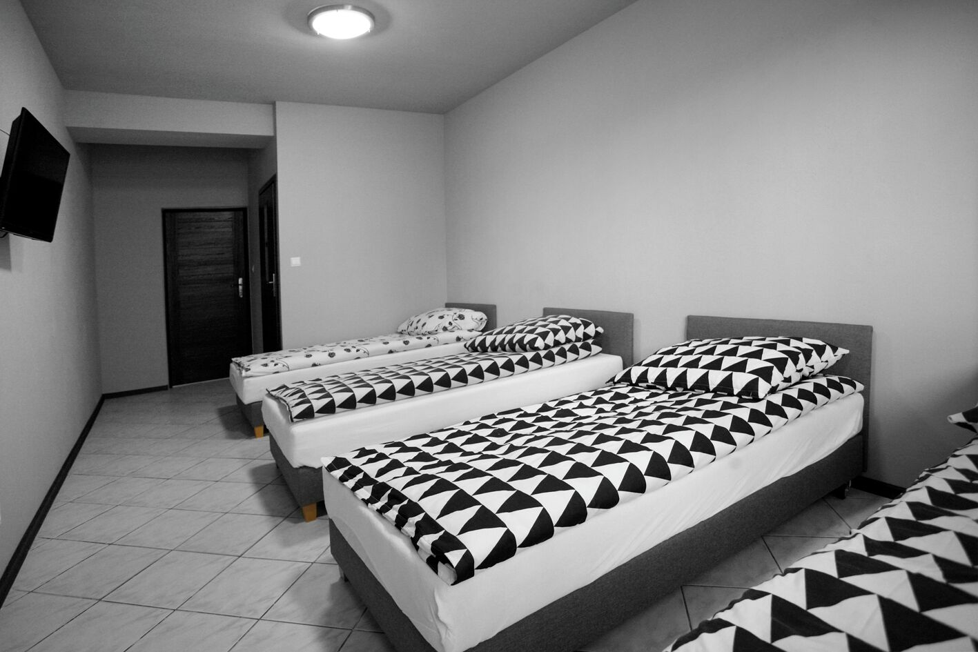 A room with three beds, each with geometric-patterned bedding, a flat-screen TV on the wall, and a tiled floor.