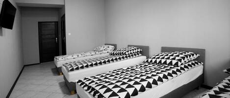 A room with three beds, each with geometric-patterned bedding, a flat-screen TV on the wall, and a tiled floor.
