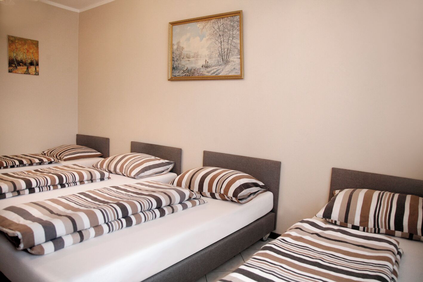 A hotel room with three beds, each with striped bedding, a painting on the wall, and a small painting on the left.