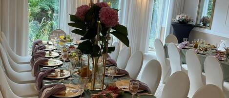 A formal dining room with a long table set for a meal, surrounded by white chairs, and a chandelier above.