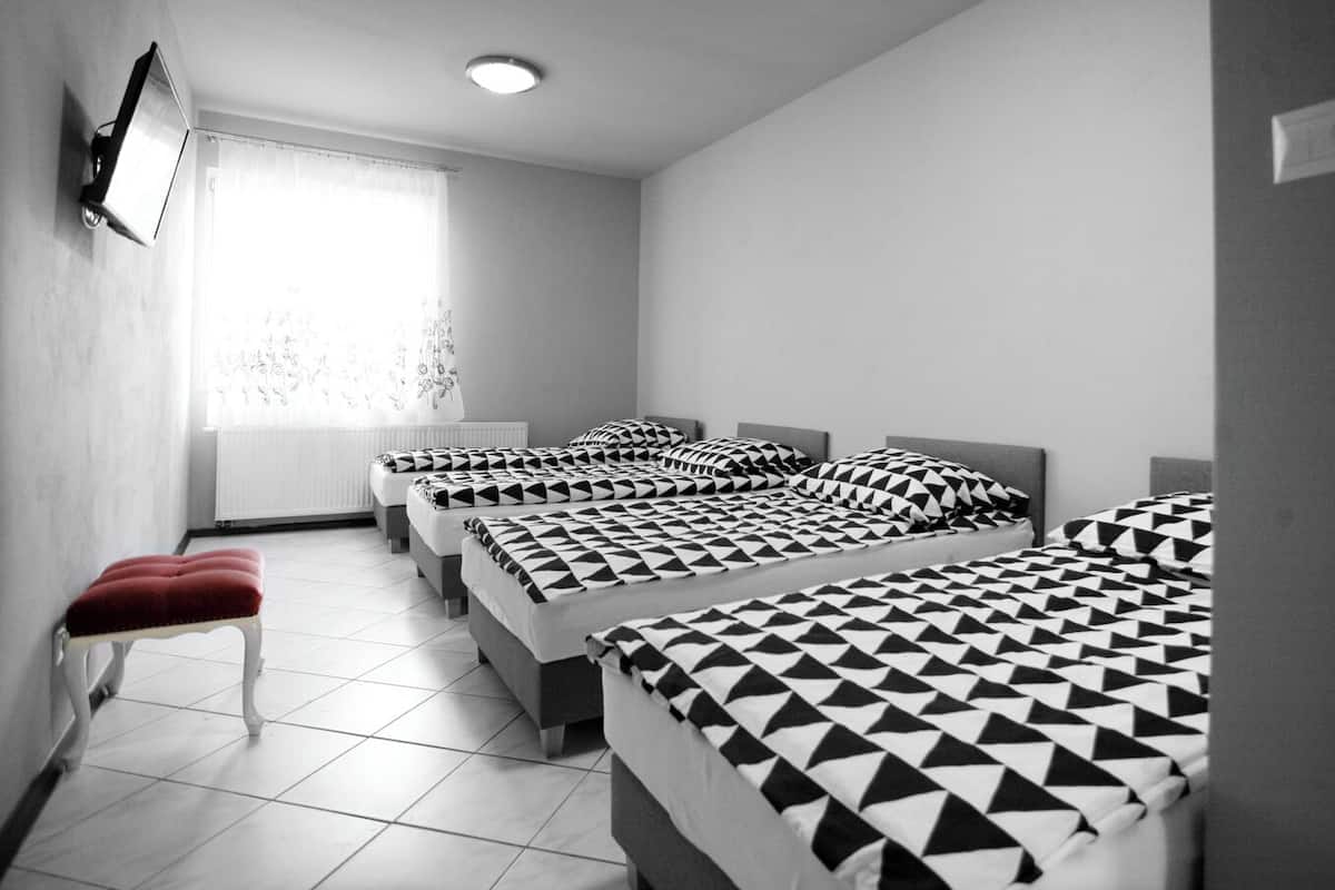 Une chambre d’hôtel avec quatre lits disposés en rangée, un banc rouge, un téléviseur fixé au mur et une fenêtre avec des rideaux.