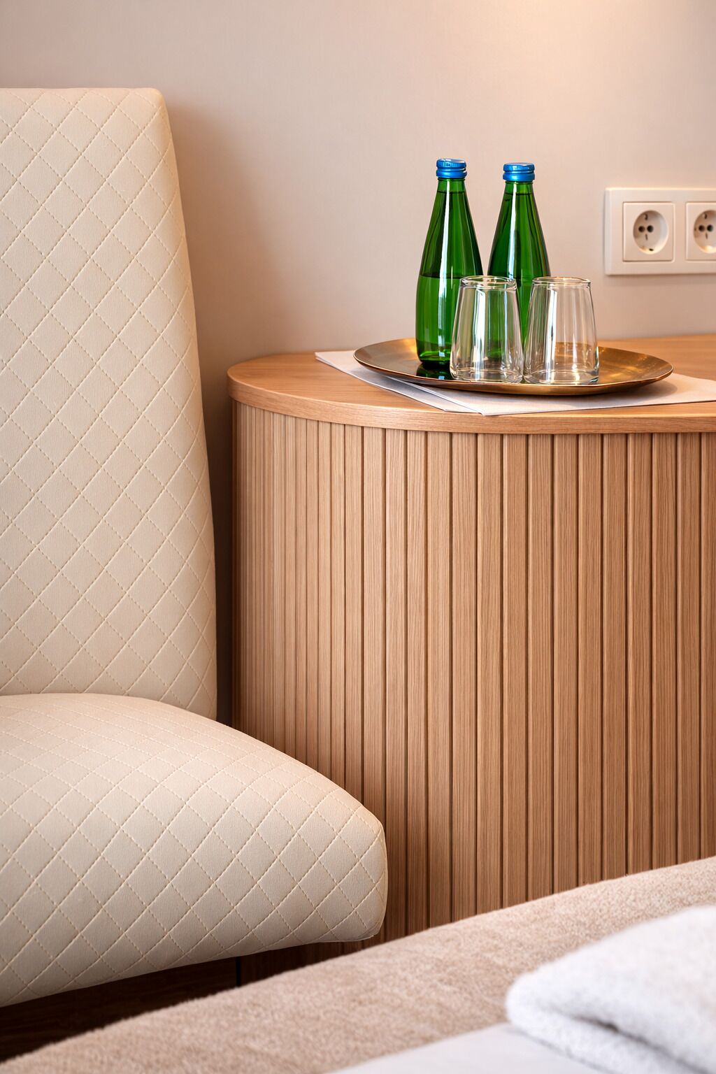 Habitación de hotel con un sillón acolchado, una mesita de madera con una bandeja que contiene dos botellas verdes y tres vasos, y un enchufe en la pared.