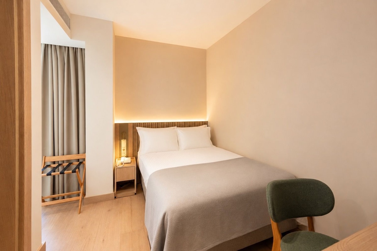 Una camera d'albergo con un letto, un comodino, una lampada, una sedia e una finestra con tende.