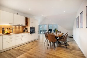 Una cocina moderna con armarios blancos, una zona de comedor con mesa y sillas de madera, y ventanas grandes.