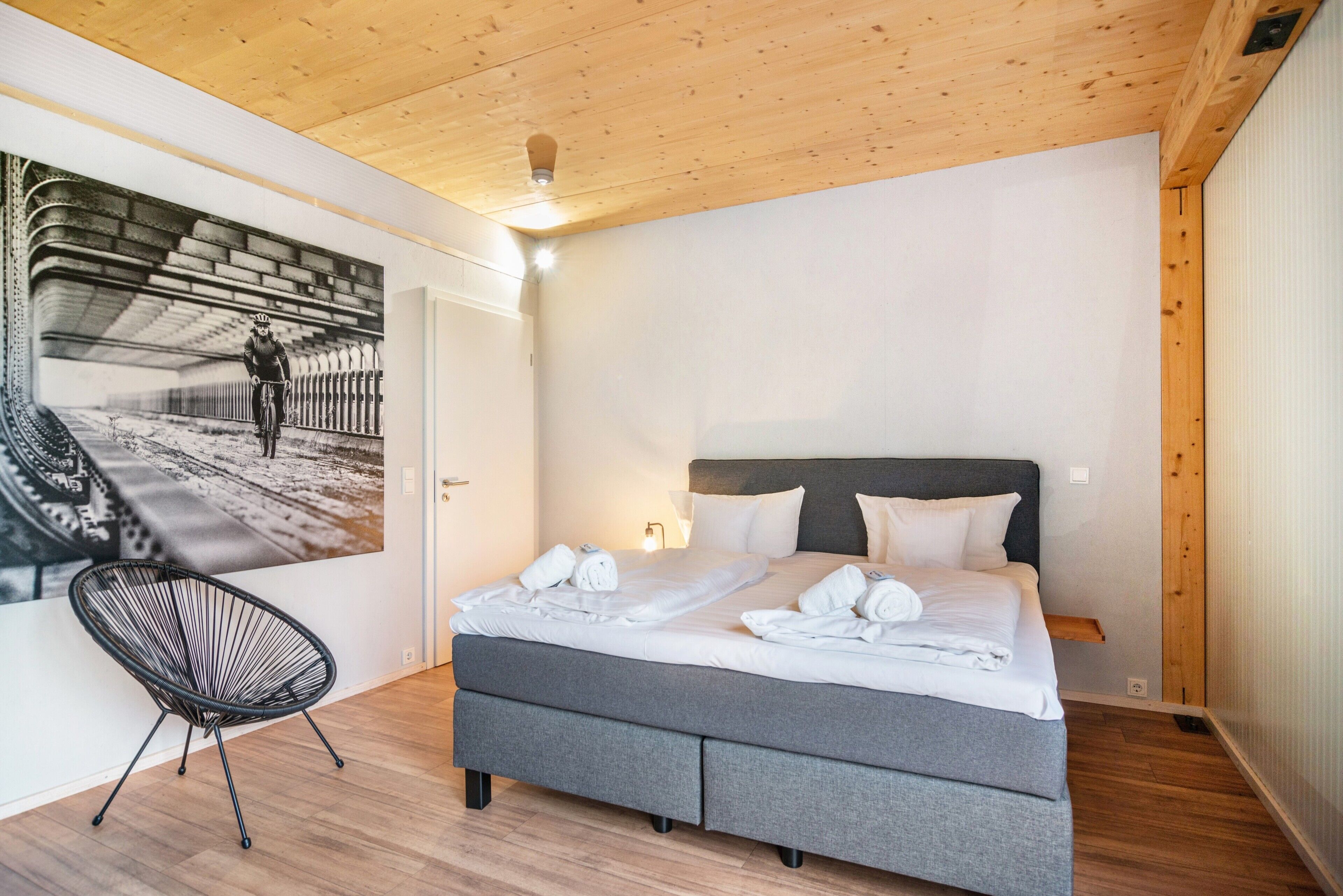 A bedroom with a bed, a chair, a wall mural, and a wooden ceiling.