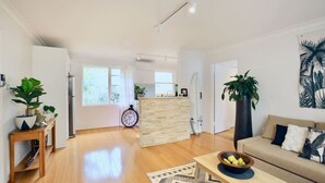 Un salón amplio con suelo de madera, un sofá, una mesa de centro y una zona de cocina con encimera y una tabla de surf.