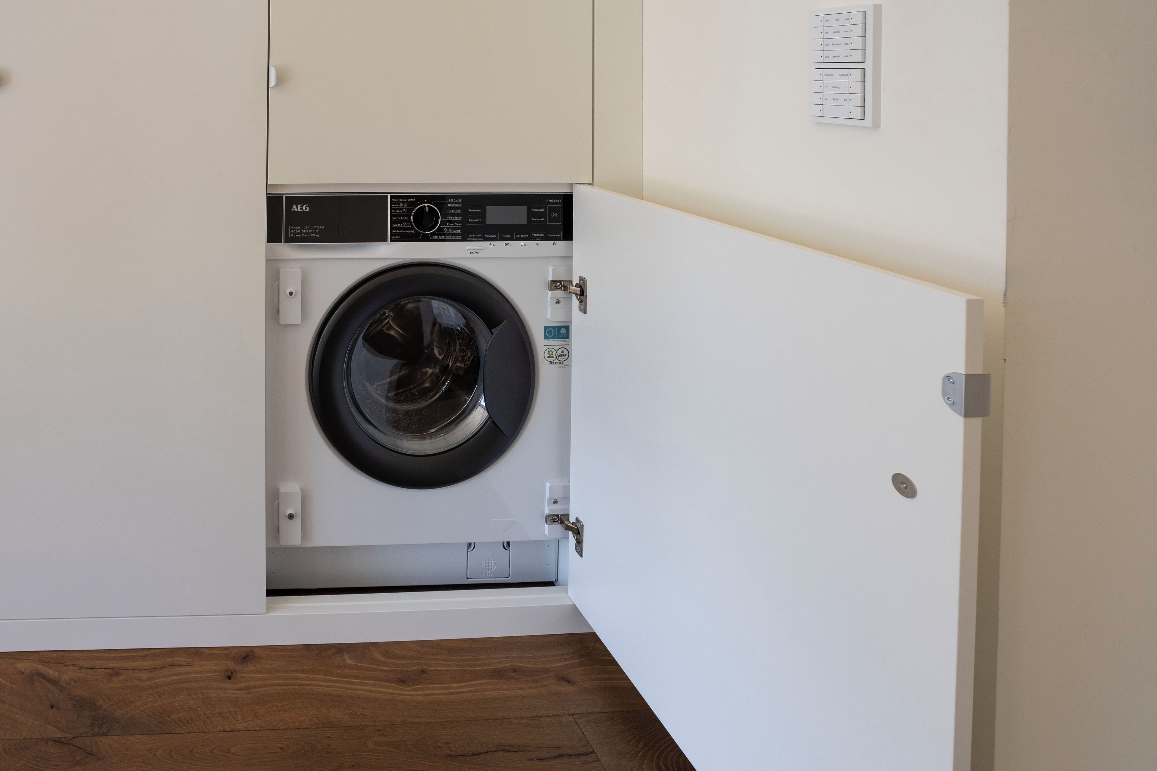 A compact, modern laundry room with a front-loading washing machine, a control panel, and a wall-mounted electrical outlet.