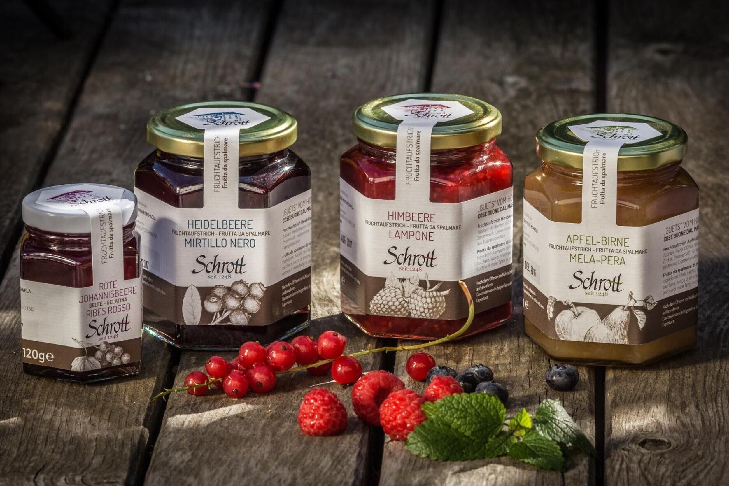Four jars of Schrott brand jam on a wooden surface with berries and mint leaves.