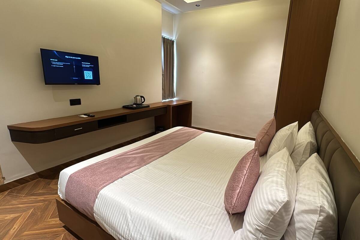 A modern hotel room with a large bed, a flat-screen TV mounted on the wall, a small table with a kettle, and a window with curtains.