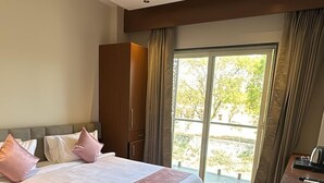 Hotelkamer met een groot bed, een plafondventilator, een houten vloer, een bureau met waterkoker en uitzicht op groen buiten.