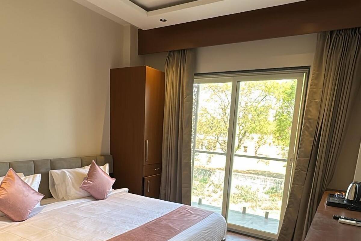 A hotel room with a large bed, a ceiling fan, a wooden floor, a desk with a kettle, and a view of greenery outside.