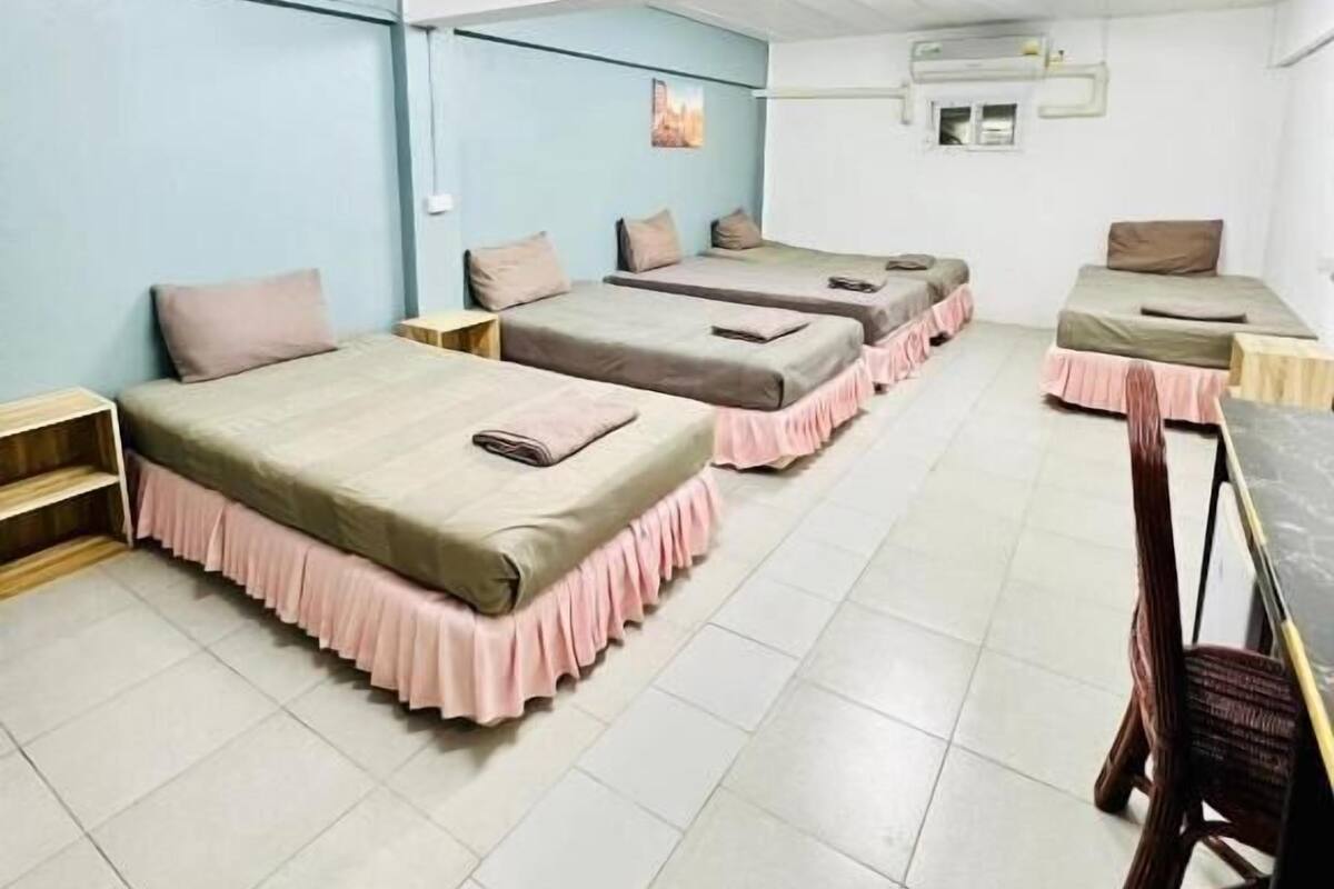 A room with three beds, each with a pink skirted bedspread, a wooden nightstand, and a wall-mounted air conditioning unit.