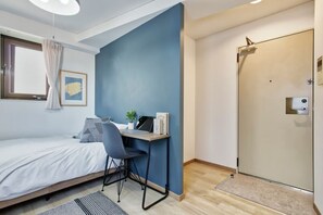 Ein Schlafzimmer mit einem Bett, einem Schreibtisch mit Laptop, einem Sessel und einer Tür.