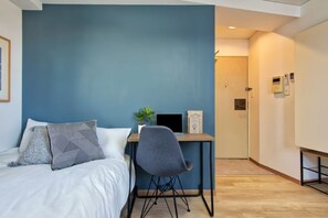 Ein Schlafzimmer mit einem Bett, einem Schreibtisch und einem Stuhl. Im Hintergrund ist eine Tür zu sehen.