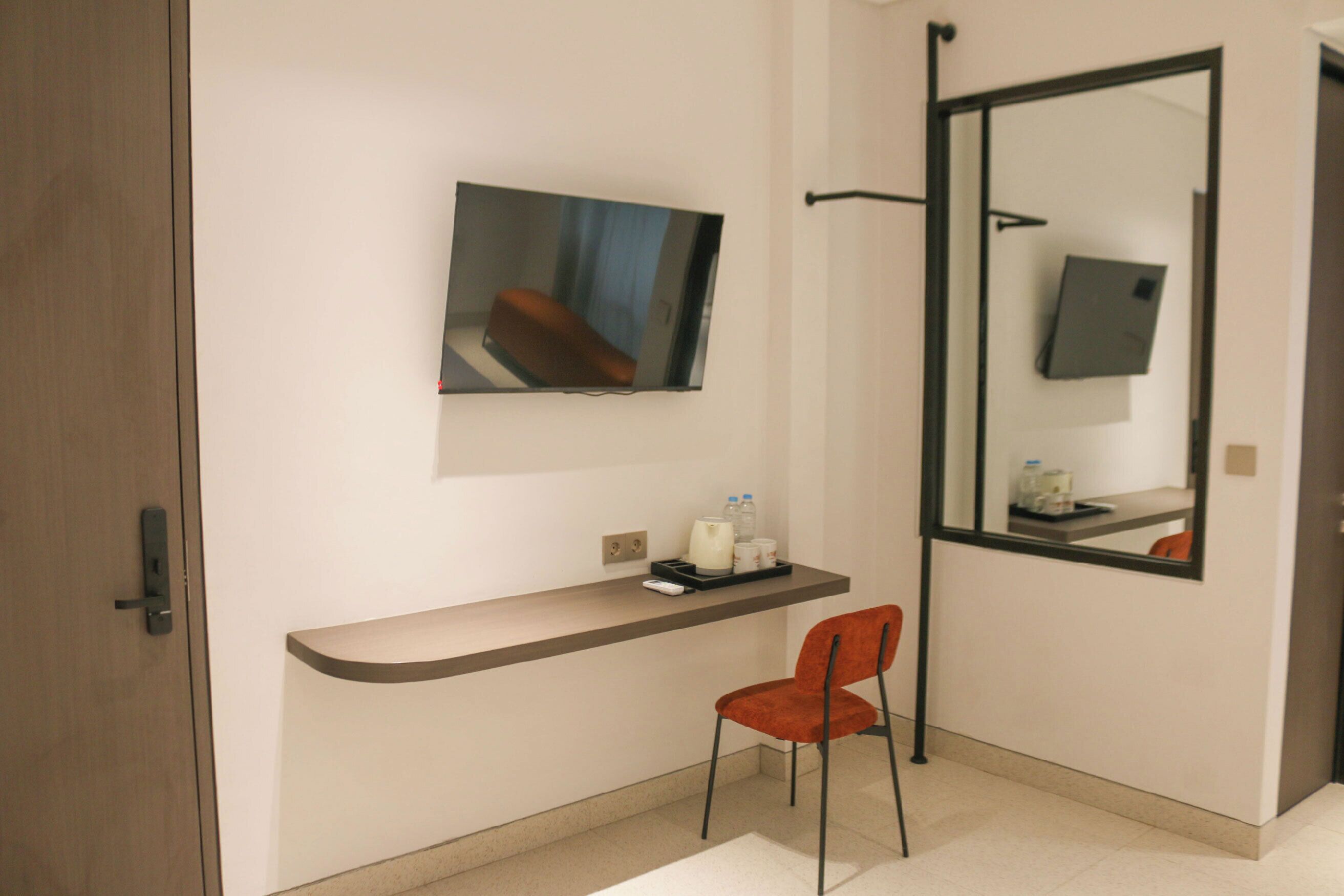 Quarto de hotel moderno com televisão de tela plana na parede, uma pequena escrivaninha com cadeira e um espelho grande refletindo o quarto.