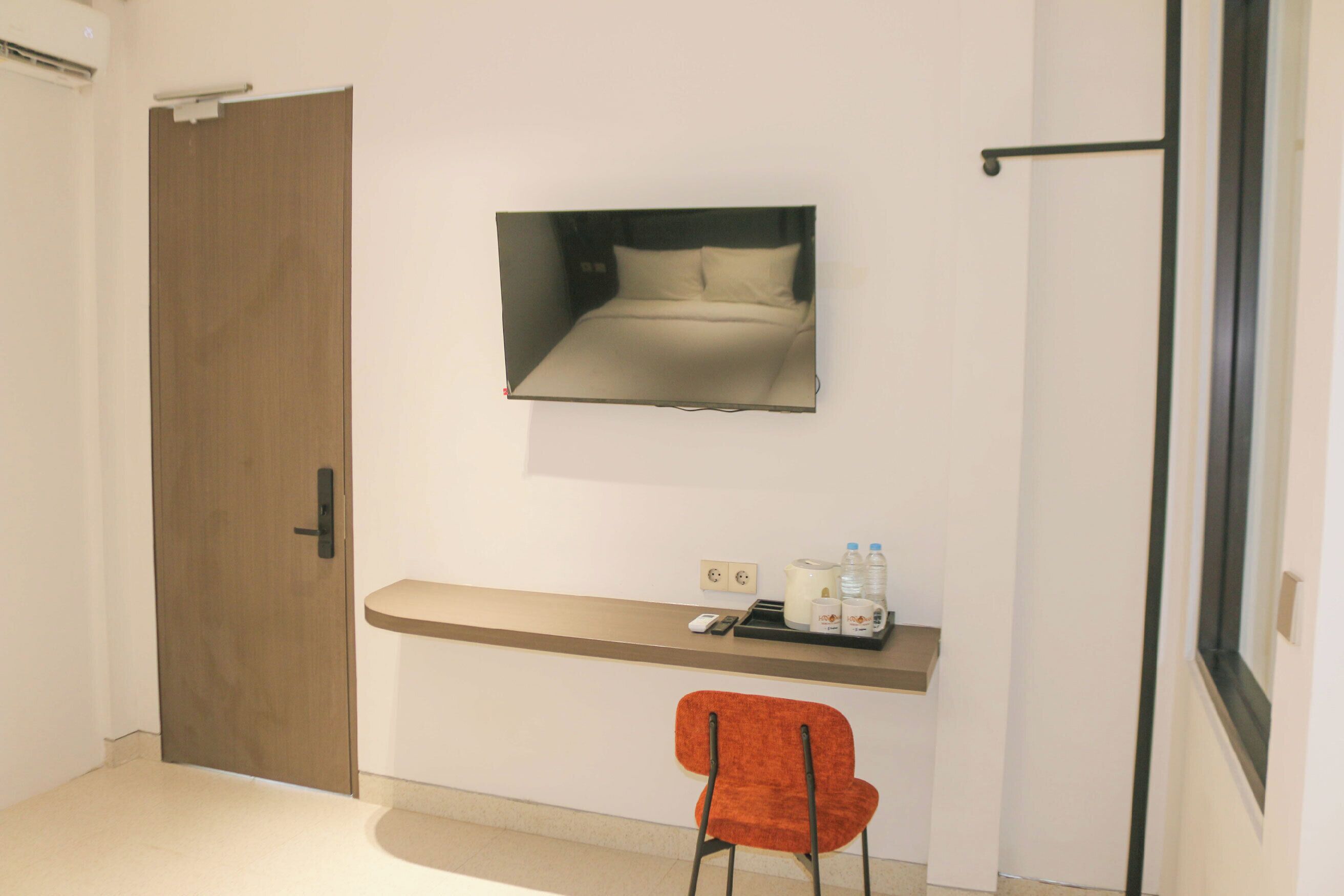 A hotel room with a wooden door, a bed with a headboard, a small desk with a chair, and a mirror on the wall.