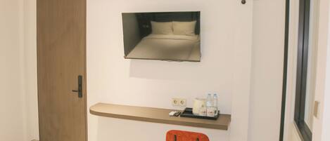 A hotel room with a wooden door, a bed with a headboard, a small desk with a chair, and a mirror on the wall.