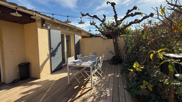 Eine Holzterrasse mit Tisch und Stühlen, ein überdachter Bereich mit Decke und ein Baum mit wenigen Blättern.