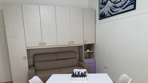 Um espaço compacto para viver, com uma mesa de jantar branca, quatro cadeiras brancas, um sofá bege e uma pequena estante com objetos decorativos.