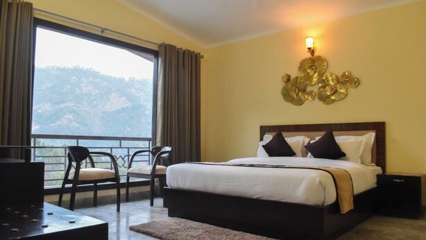 A hotel room with a large bed, a desk, and a chair. There is a balcony with a view of the mountains. - Pinewood Manor Kasauli (Solan)