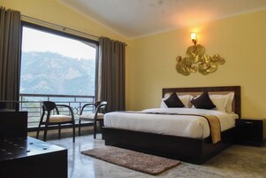 A hotel room with a large bed, a desk, and a chair. There is a balcony with a view of the mountains.