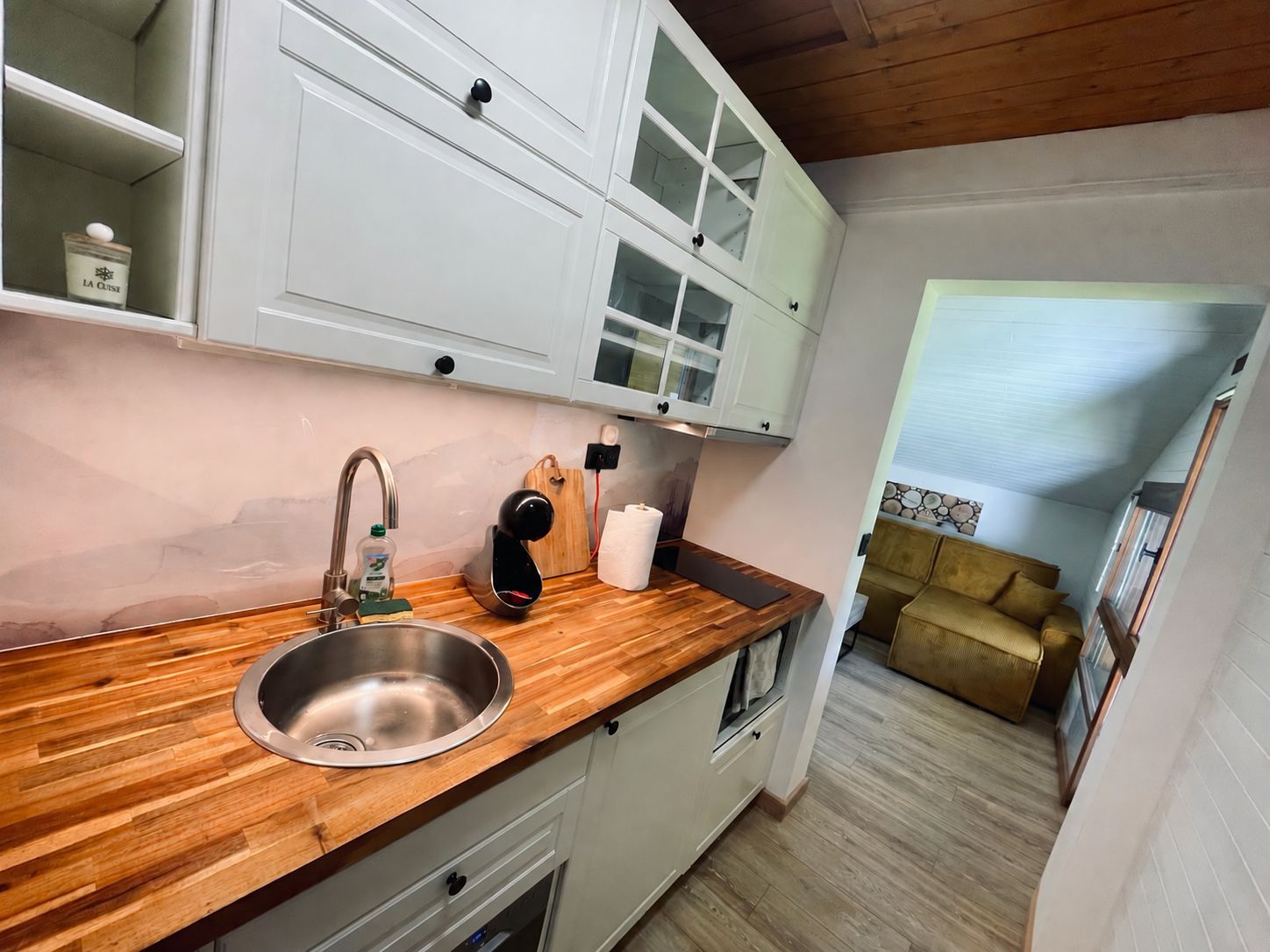 A kitchen with a wooden countertop, stainless steel sink, white cabinets, and a brown sofa in the adjacent living area.