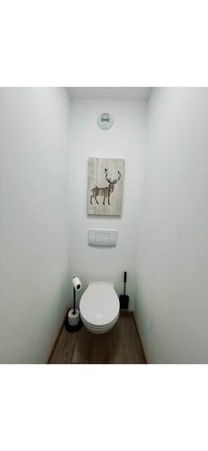 Un baño pequeño con inodoro blanco, suelo de madera, una lámpara de pared y una obra de arte de madera con un ciervo.