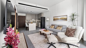 Ein modernes Wohnzimmer mit einem grauen Sofa, einem Couchtisch und einem kleinen Beistelltisch. Im Hintergrund ist eine Küche mit einem schwarzen Kühlschrank und einem Essbereich zu sehen.