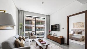 Ein modernes Wohnzimmer mit einem grauen Sofa, einem Couchtisch und einem Fernseher. Ein großes Fenster bietet Blick auf eine Baustelle.